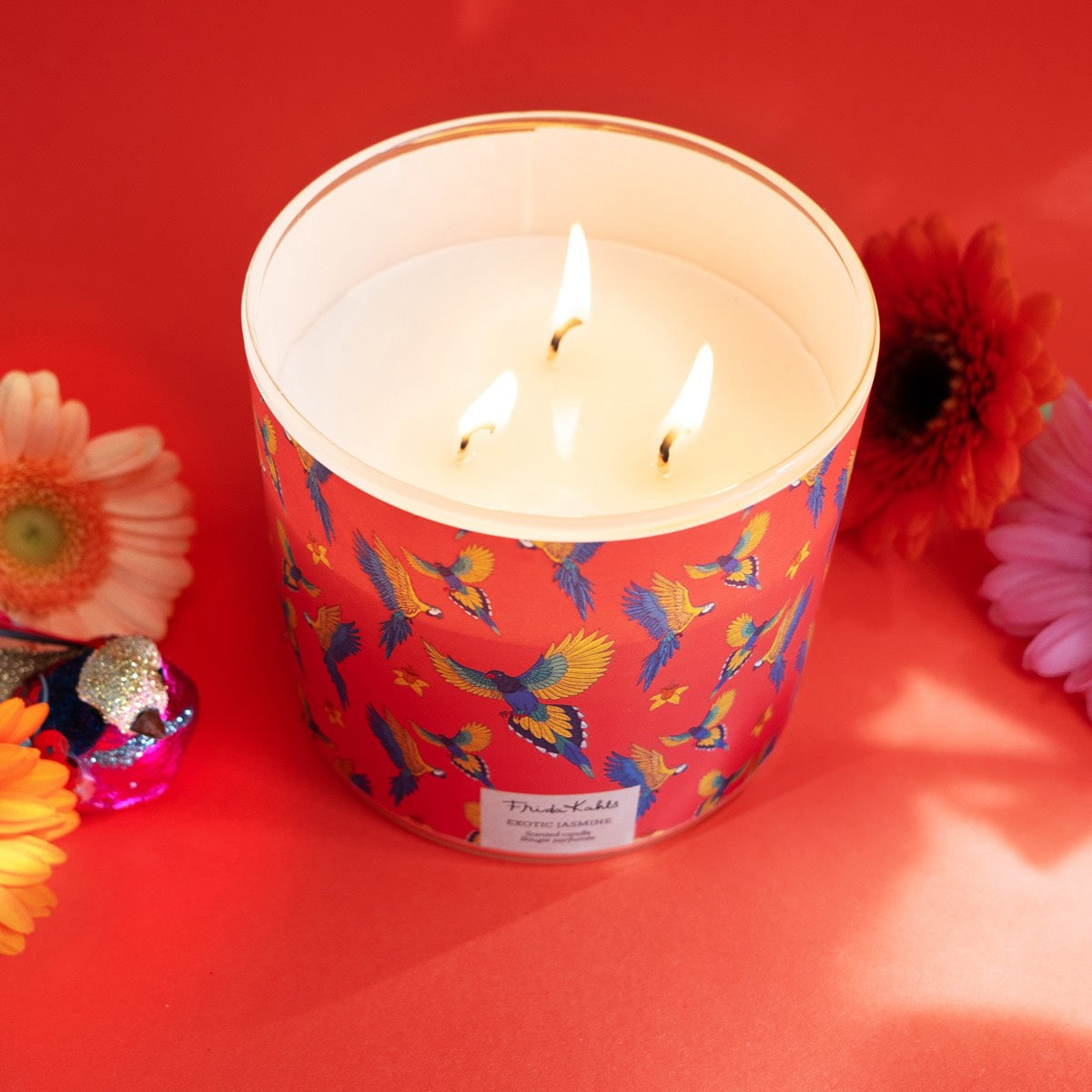 Frida Kahlo Duftkerze ‚Exotic Jasmine‘ (400g) – Moodbild der offiziellen Lizenzkerze im blumig-künstlerischen Design. Die detailverzierte Duftkerze steht frontal auf einem orangenem Hintergrund, der Deckel wurde entfernt und die drei Baumwolldochte brennen – ein stilvolles Wohnaccessoire und eine besondere Geschenkidee für Duftkerzen-Liebhaber.