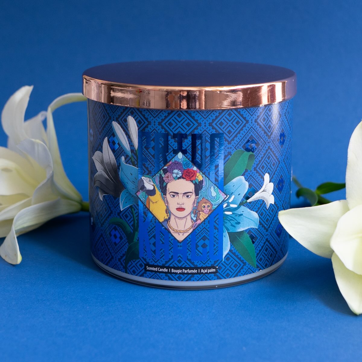 Frida Kahlo Duftkerze ‚Frida‘ (400g) – Moodbild der offiziellen Lizenzkerze  im blauem Design und mit einem Frida Kahlo Porträt. Die kunstvoll verzierte Kerze steht frontal auf einer blauen Unterlage und links sowie rechts von ihr liegen zwei weiße Blüten – dekoratives Wohnaccessoire und ideale Geschenkidee für Duftkerzen-Liebhaber.