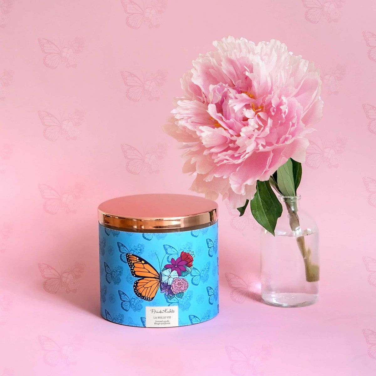 Frida Kahlo Duftkerze ‚La belle Vie‘ (400g) – Moodbild der offiziellen Lizenzkerze im frühlingshaften Design. Die kunstvoll verzierte Kerze steht frontal auf einer rosafarbenen Unterlage. Rechts von ihr steht eine rosa Blume in einer Vase – dekoratives Wohnaccessoire und ideale Geschenkidee für Duftkerzen-Liebhaber.