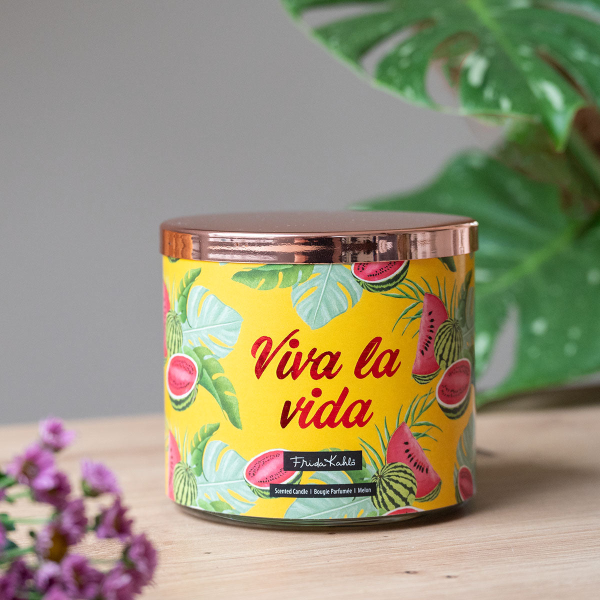 Frida Kahlo Duftkerze ‚Viva la Vida‘ (400g) – Moodbild der offiziellen Lizenzkerze  im sommerlichen Design. Die kunstvoll verzierte Kerze steht frontal auf einem Holztisch. Im Vordergrund ist eine violette Blüte angeschnitten und im Hintergrund große grüne Blätter  – dekoratives Wohnaccessoire und ideale Geschenkidee für Duftkerzen-Liebhaber.