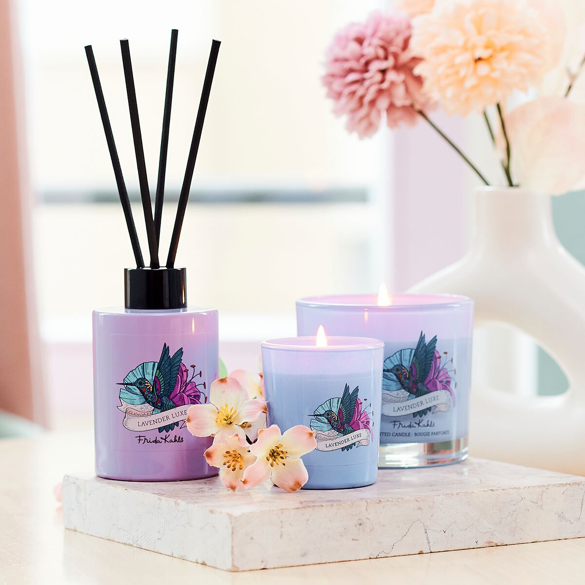 Frida Kahlo Lavender Luxe Kollektion Duftstäbchen zusammen mit Duftkerzen. Stilvoll angerichtet auf einem Mamorstein mit weißen Blüten. Perfekte Raumerfrischung, die eine elegante und hochwertige Dekoration darstellen.