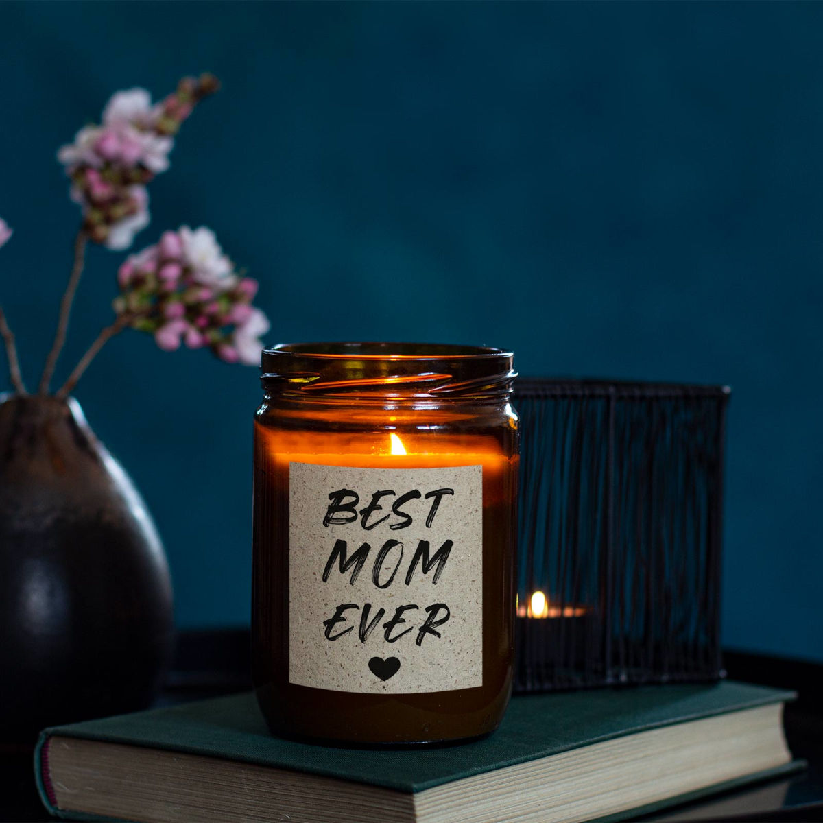 Moodbild: Handgemachte ‚Best Mom ever‘ Duftkerze von mad candle aus Sojawachs mit beschriftetem Etikett – dekoriert auf einem Buch mit einem schwarzen Teelichhalter und einer Vase im Hintergrund. Edles Geschenk für besondere Anlässe oder stilvolle Home-Deko.
