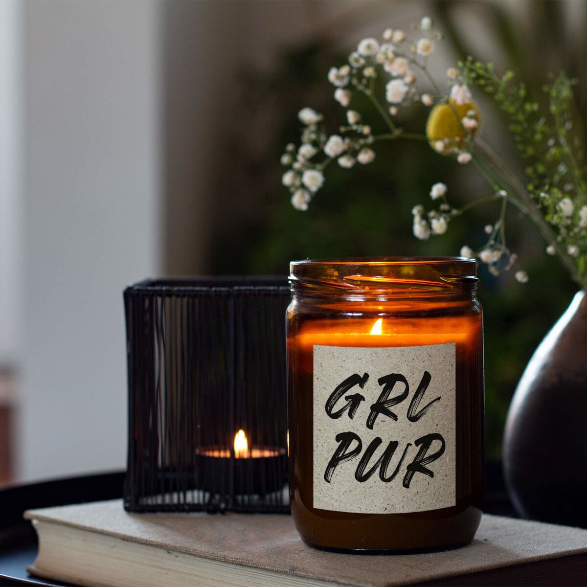 Moodbild: Handgemachte ‚ Girlpower‘ Duftkerze von mad candle aus Sojawachs mit beschriftetem Etikett – dekoriert auf einem Buch mit einem schwarzen Teelichthalter und Blumenvase im Hintergrund. Edles Geschenk für besondere Anlässe oder stilvolle Home-Deko.