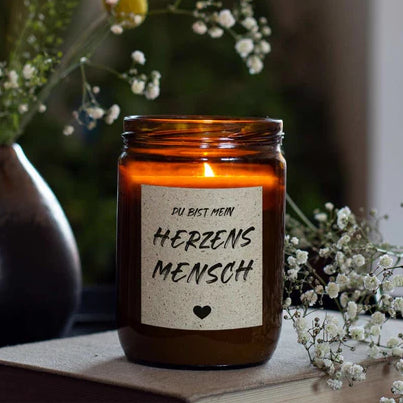 mad candle Duftkerze aus Sojawachs mit dekorativen Etikett, Teelicht und Blume im Hintergrund. Die Kerze brennt und steht auf einem Buch.