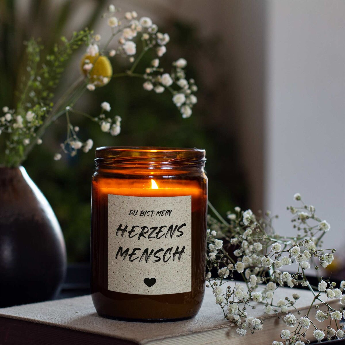 mad candle Duftkerze aus Sojawachs mit dekorativen Etikett, Teelicht und Blume im Hintergrund. Die Kerze brennt und steht auf einem Buch.