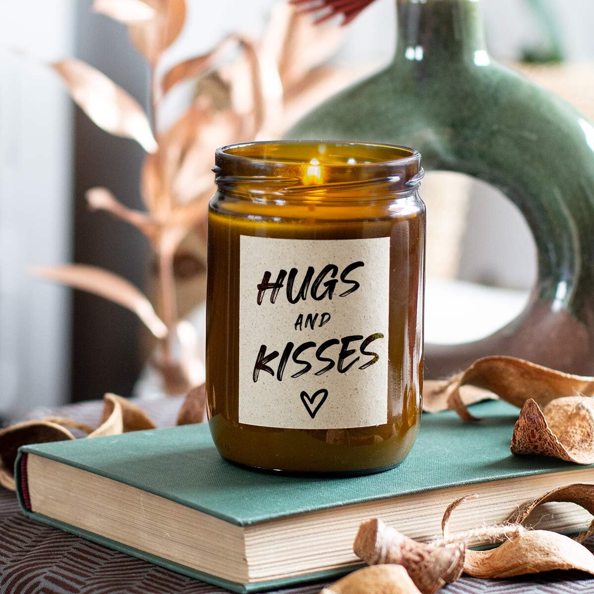 Moodbild: Handgemachte ‚Hugs and Kisses‘ Duftkerze von mad candle aus Sojawachs mit beschriftetem Etikett – dekoriert auf einem Buch mit getrockneten Blättern und einer Vase im Hintergrund. Edles Geschenk für besondere Anlässe oder stilvolle Home-Deko.