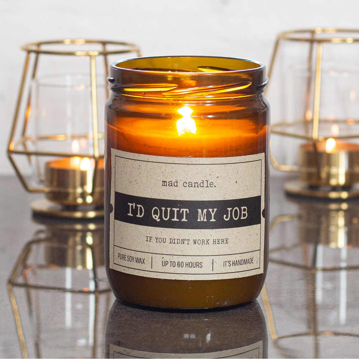Moodbild: Handgemachte ‚I'd quit my Job‘ Duftkerze von mad candle aus Sojawachs mit beschriftetem Etikett – dekoriert auf einem Tisch mit zwei goldenen Teelichthaltern im Hintergrund. Edles Geschenk für Kollegen oder stilvolle Büro-Deko.