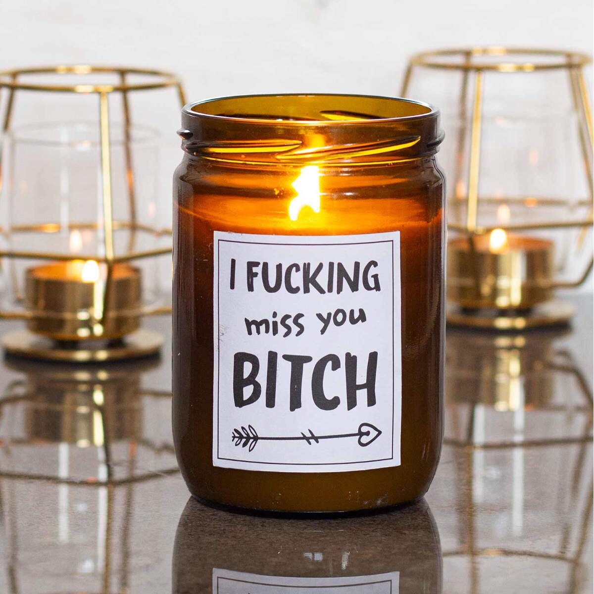 Moodbild: Handgemachte ‚I fucking miss you bitch‘ Duftkerze von mad candle aus Sojawachs mit beschriftetem Etikett – dekoriert auf einem Tisch mit zwei goldenen Teelichthaltern im Hintergrund. Stilvolles Geschenk für deine beste Freundin oder edle Home-Deko.