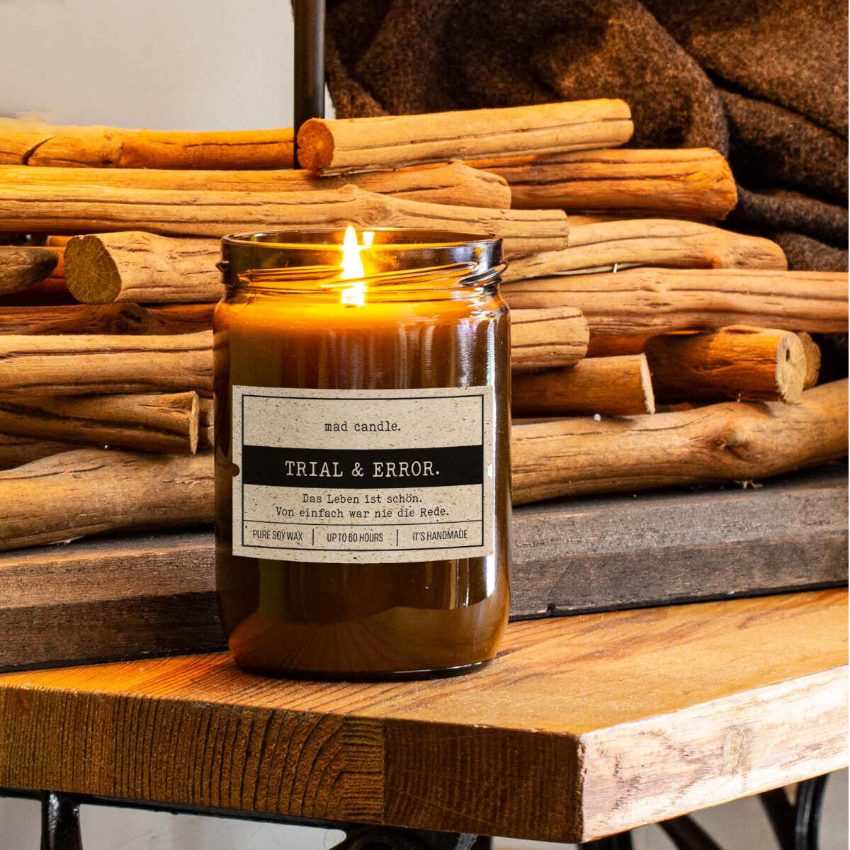 Handgemachte ‚Trail & Error.‘ Duftkerze von mad candle aus Sojawachs mit beschriftetem Etikett – dekoriert auf einem Holztisch mit Holzstücken im Hintergrund. Edles Geschenk für besondere Anlässe oder stilvolle Home-Deko.