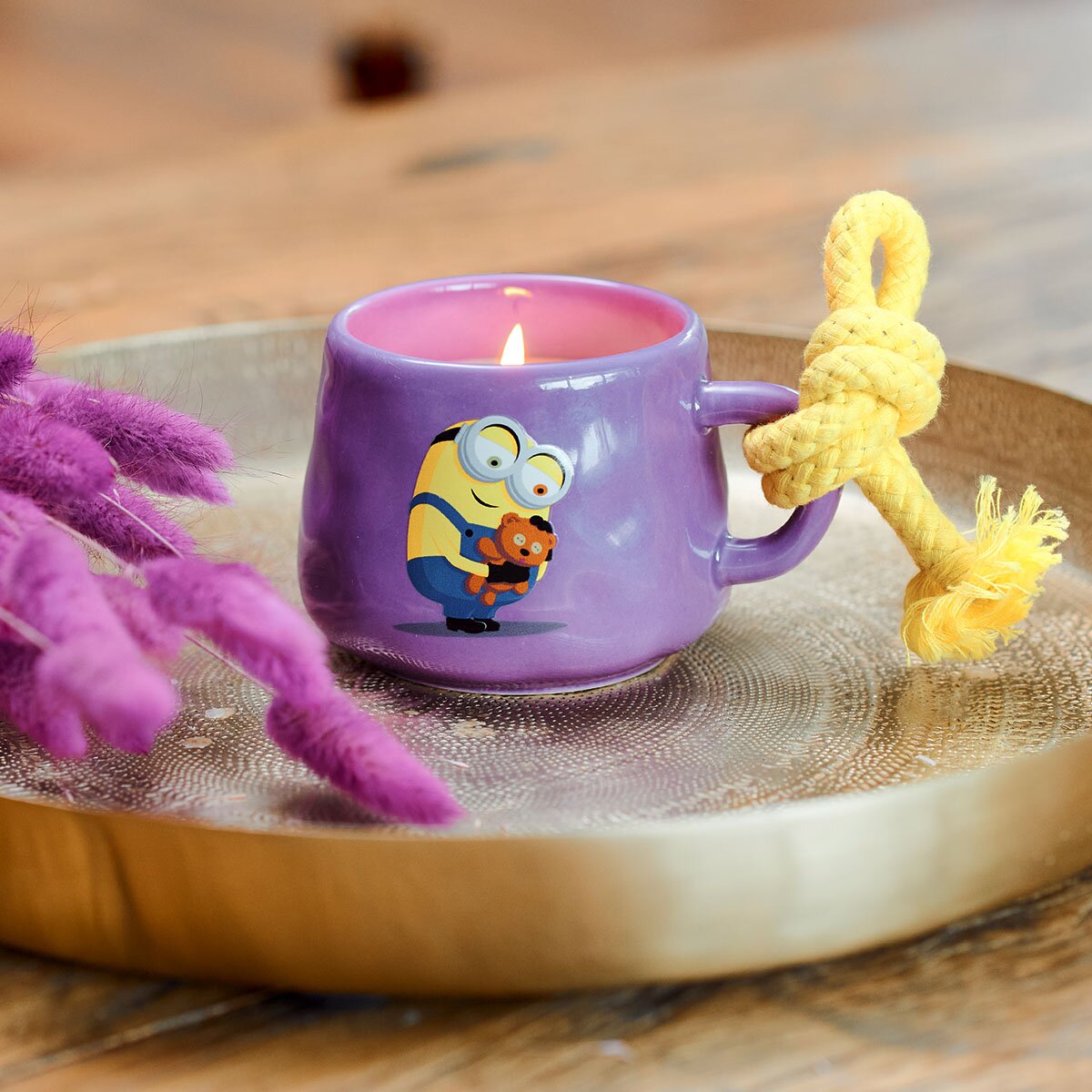 Minions Duftkerze Bob & Tim (200g) in lila Keramiktasse mit gelber Kordel. Als Motiv auf der Tasse ist Bob abgebildete, der sein Kuschelbären Tim umarmt. Der Docht brennt und die Kerze befindet sich auf einem hochwertigen Holzteller zusammen mit lila Gräser.