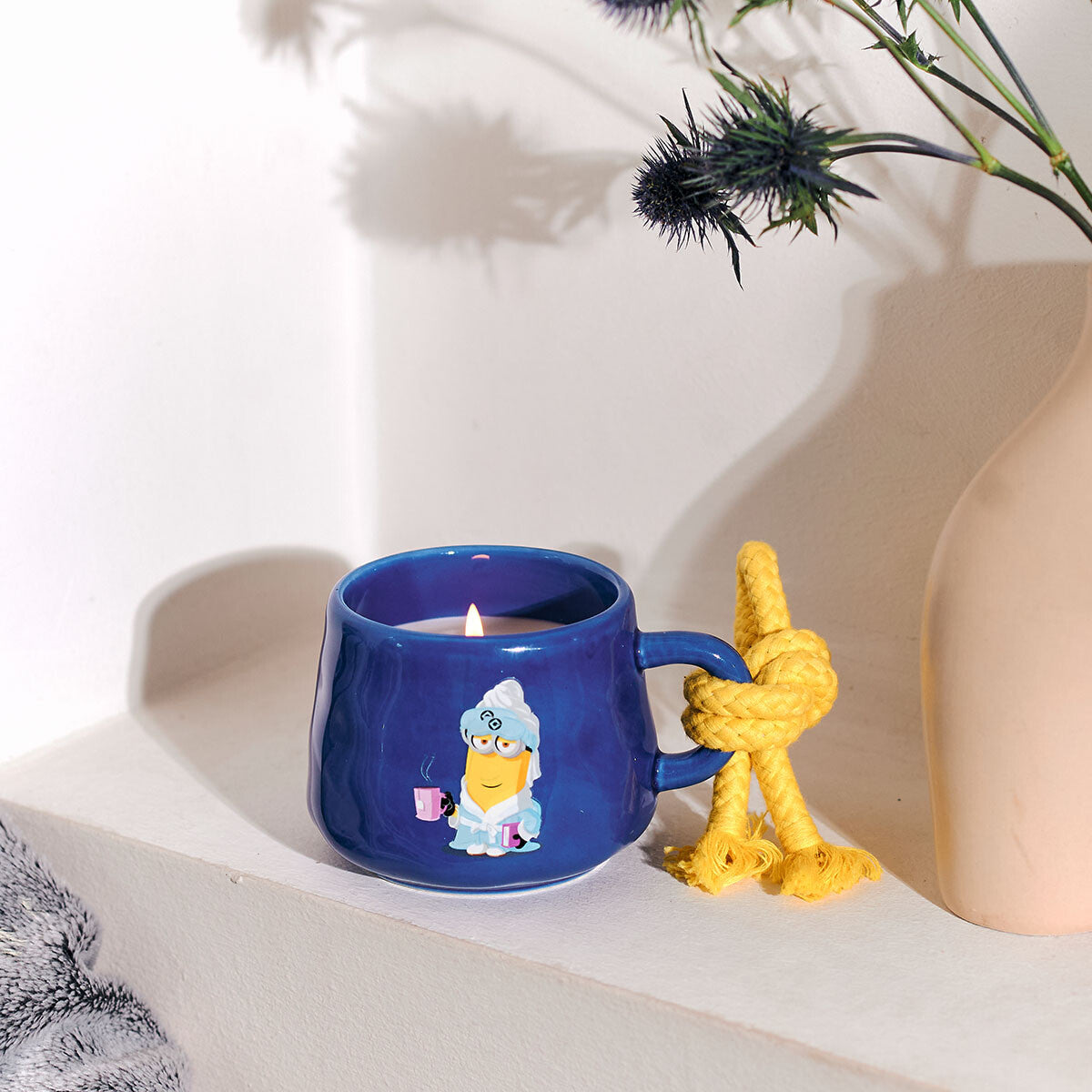 Minions Duftkerze Kevin (200g) in blauer Keramiktasse mit gelber Kordel. Als Motiv auf der Tasse ist Kevin im Bademantel abgebildete, der ein Tee und ein Buch in der Hand hält. Der Docht brennt und die Kerze befindet sich in einem gemütlichen Wohnzimmer. Neben ihr steht eine Distel.