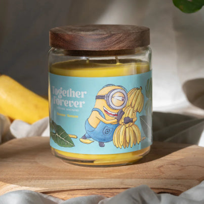 Minions XXL-Duftkerze ‚Together Forever‘ (510g) – Moodbild der offiziellen Lizenzkerze mit farbenfrohem Minions-Motiv, Palmenblättern und einer Bananenstaude. Vegane Duftkerze im Glas als stylische Deko oder Geschenk für Fans.