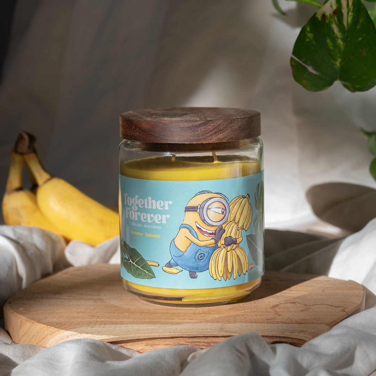 Minions XXL-Duftkerze ‚Together Forever‘ (510g) – Moodbild der offiziellen Lizenzkerze mit farbenfrohem Minions-Motiv, Palmenblättern und einer Bananenstaude. Vegane Duftkerze im Glas als stylische Deko oder Geschenk für Fans.