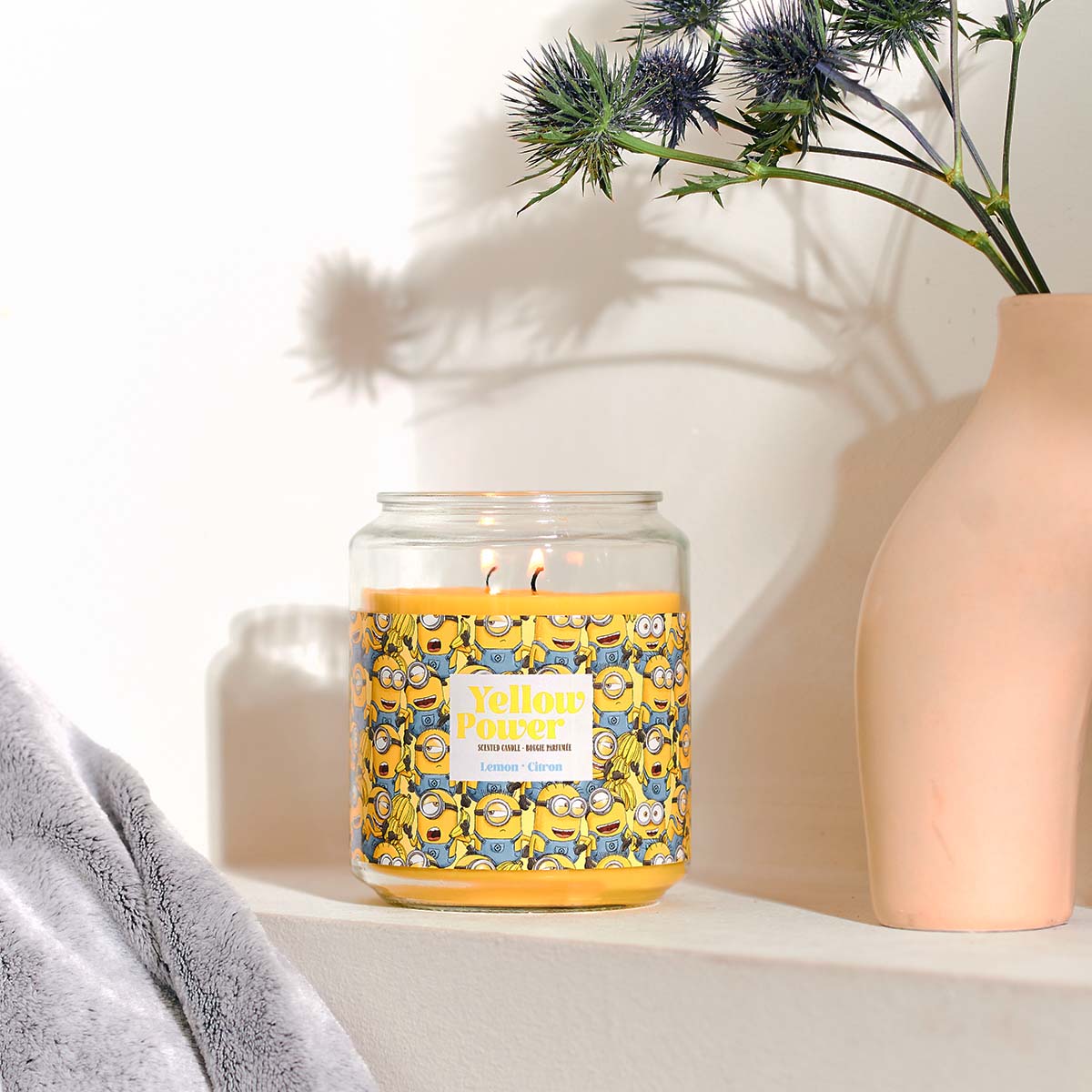 Minions XXL-Duftkerze ‚Yellow Power‘ (510g) – Moodbild der offiziellen Lizenzkerze mit gelbem Minions-Motiv, einer Blumenvase und einer Kuscheldecke. Vegane Duftkerze im Glas als stylische Deko oder Geschenk für Kinder.