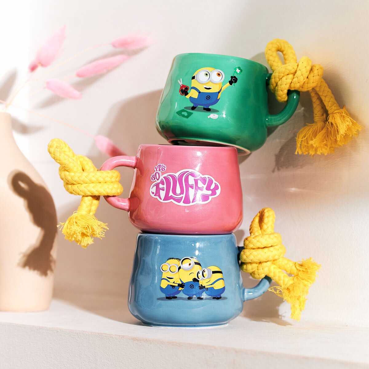 Minions Duftkerzen Bob, Fluffy und Dave, Stuart & Bob (3x 200g) in grüner, roter und blauer Keramiktasse mit gelber Kordel. Die drei Tassen sind übereinander gestapelt und lehnen an einer Wand. Neben ihnen ist eine Vase mit rosa Gräsern.
