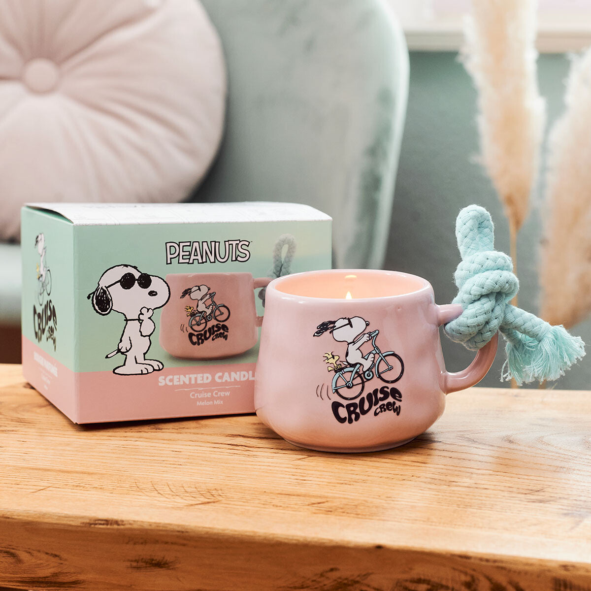 Peanuts-Duftkerze Cruise Crew (200g) in rosa Keramiktasse von Snoopy. Moodbild der Tasse mit Geschenkkarton, die in einem gemütlichen und dekorativen Wohnzimmer stehen.