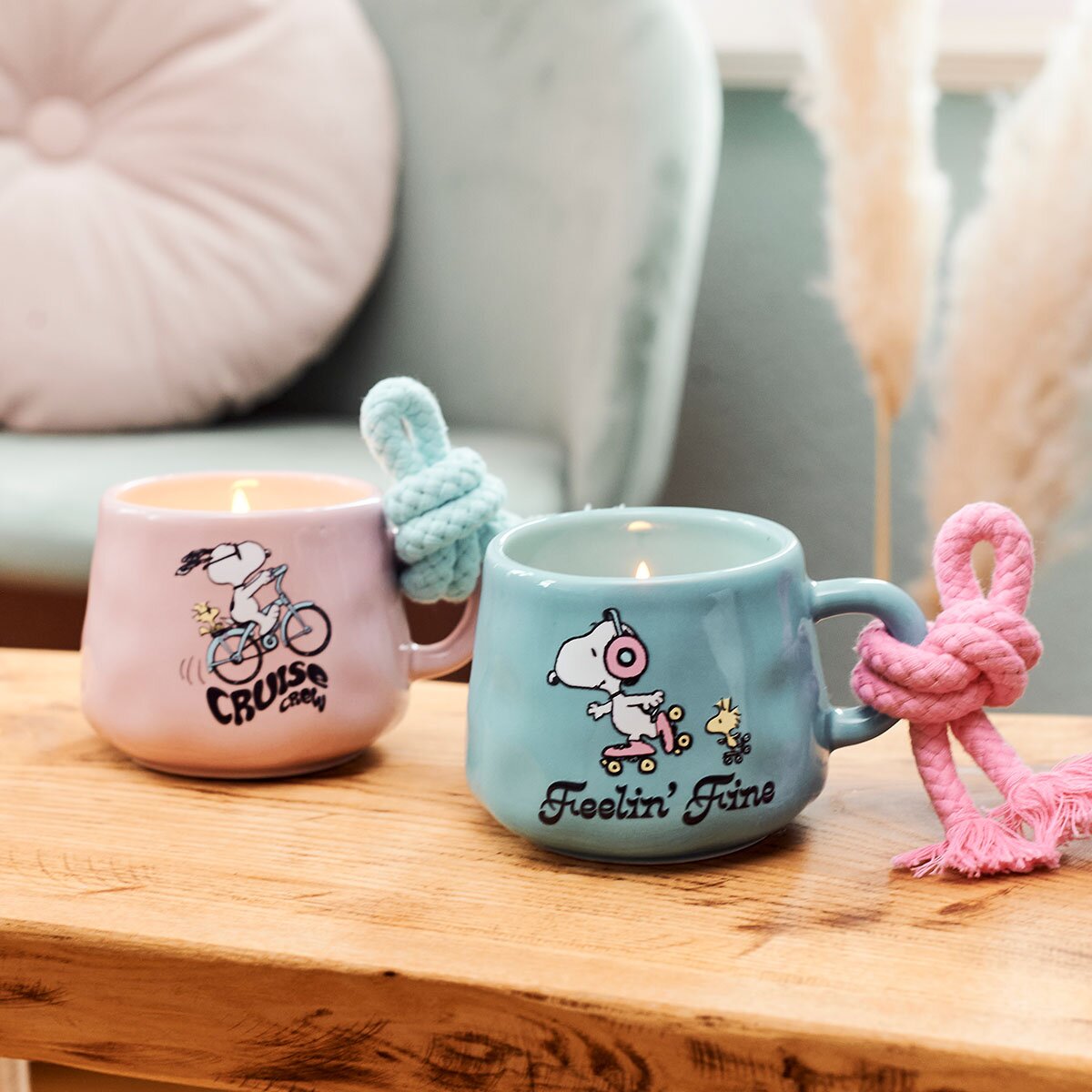 Peanuts-Duftkerzen Feeling Fine (200g) in türkisfarbener Keramiktasse und rosa Cruise Crew Keramiktasse von Snoopy. Moodbild der beiden Tassen, die in einem gemütlichen und dekorativen Wohnzimmer stehen.