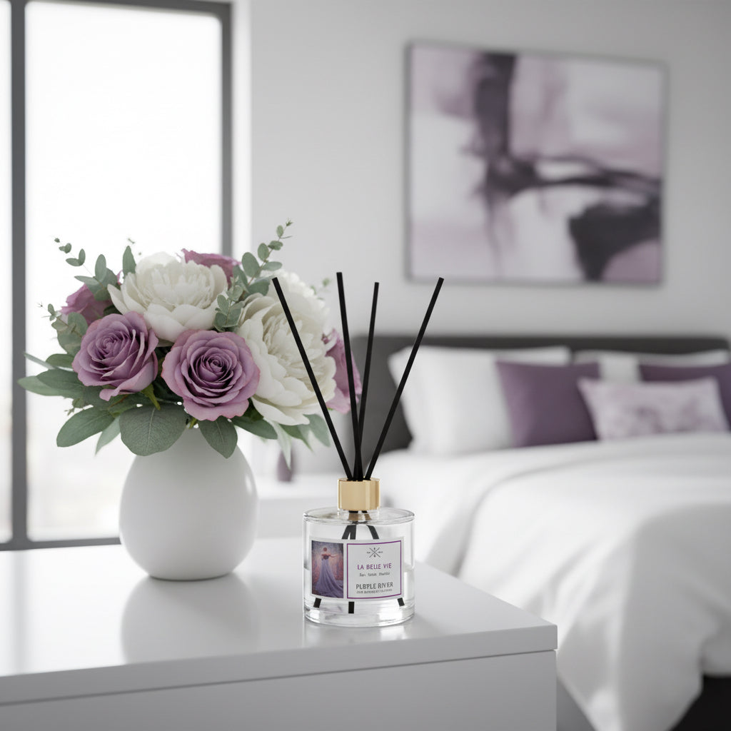 Purple River Raumduft (100 ml) mit schwarzen Schilfstäbchen und goldener Kappe auf einem Tisch im Schlafzimmer, neben einem Blumenstrauß aus lila Rosen und mit einem Bett im Hintergrund; hochwertiger Duft mit bis zu 6 Wochen Duftdauer, ideal als stilvolle Dekoration und luxuriöses Wellness-Geschenk, gefertigt in Deutschland.