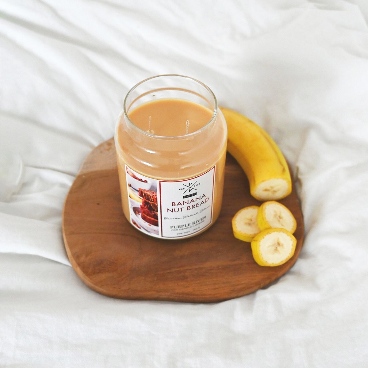 Nachhaltige Premium-Duftkerze von Purple River mit extralanger 140h-Brenndauer. Hochwertige Wellness-Deko im Glas. Made in Germany - inszeniert auf Holzbrett neben geschnittener Banane.