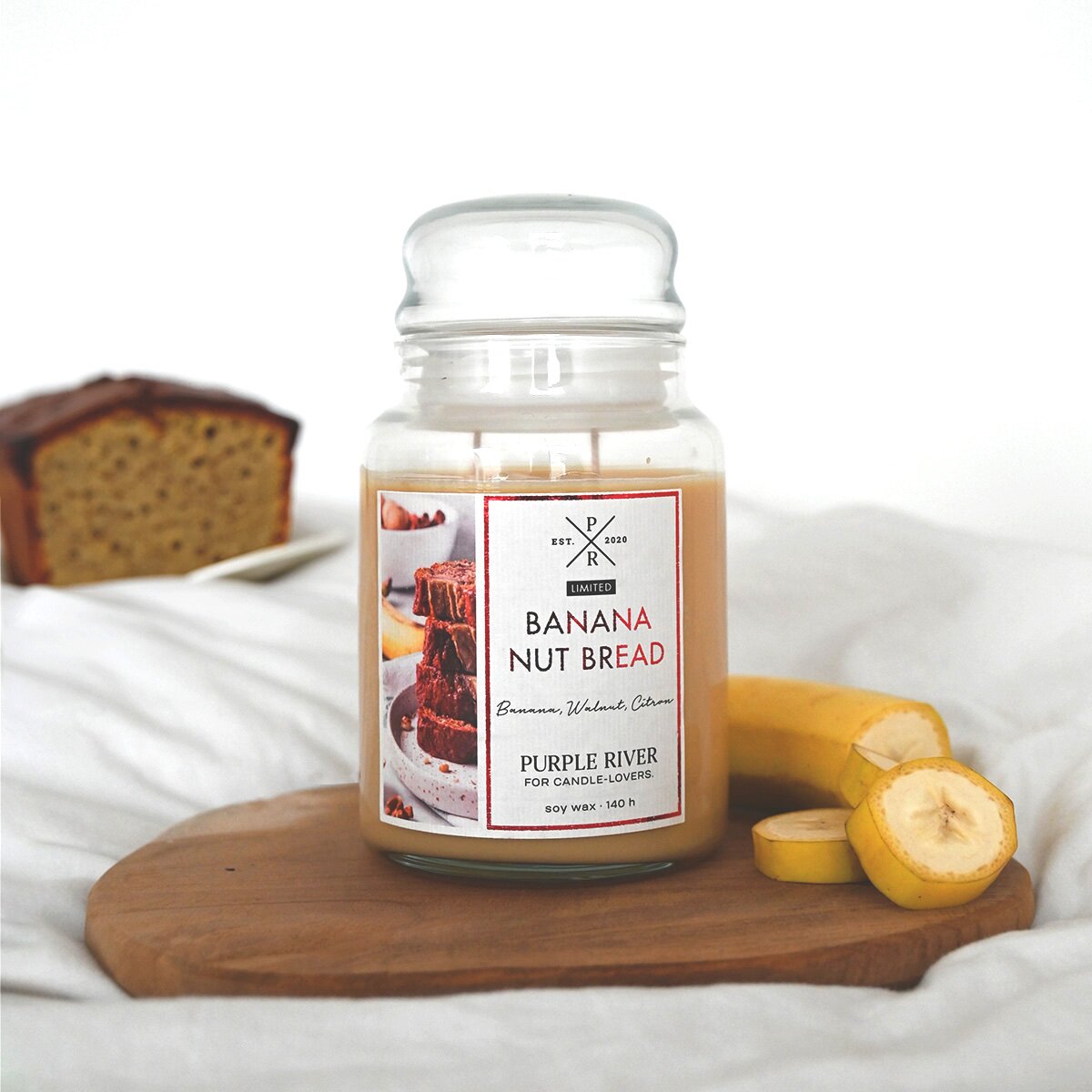 Nachhaltige Premium-Duftkerze von Purple River mit extralanger 140h-Brenndauer. Hochwertige Wellness-Deko im Glas. Made in Germany - inszeniert auf Holzbrett neben geschnittener Banane & Bananenbrot im Hintergrund. 