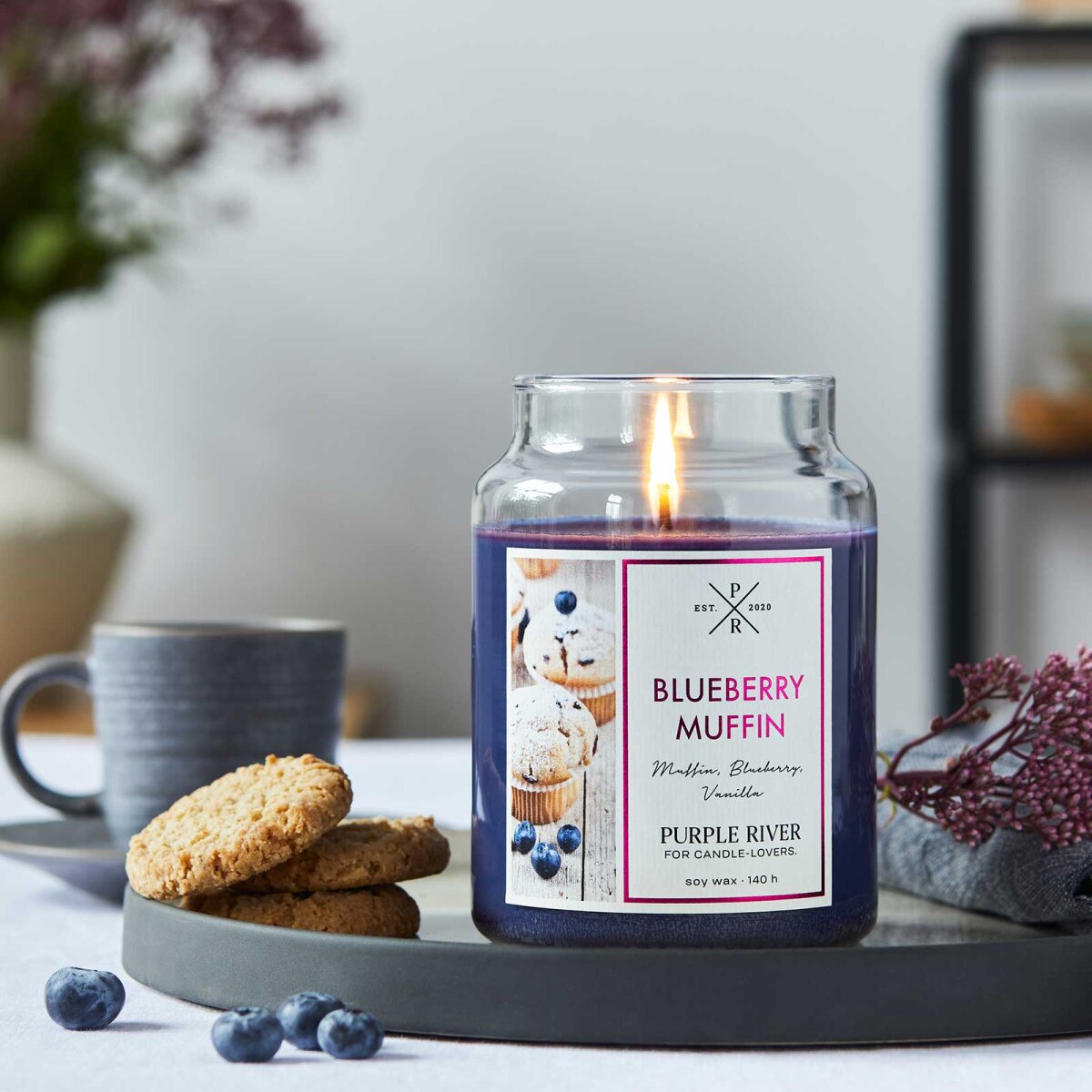 Nachhaltige Premium-Duftkerze von Purple River mit extralanger 140h-Brenndauer. Hochwertiges Geschenk im Glas. Made in Germany - inszeniert auf Keramikteller neben Plätzchen & Blaubeeren im Wohnzimmer.