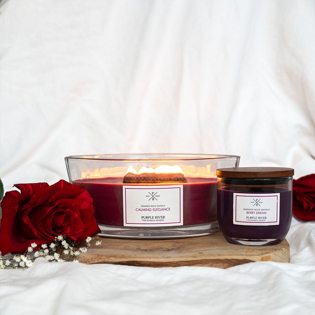 Zwei Nachhaltige Premium-Duftkerzen von Purple River in rot & lila mit langern Brenndauern. Hochwertige Wellness-Dekorationen im Glas und mit Holzdeckel. Made in Germany - inszeniert auf Holzbrett mit weißem Hintergrund und roten Rosen.