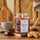 Nachhaltige Premium-Duftkerze von Purple River mit extralanger 140h-Brenndauer. Hochwertige Wellness-Deko im Glas. Made in Germany - inszeniert auf Holzbrett neben Keks, Schokolade & Keramiktasse in Küche. 