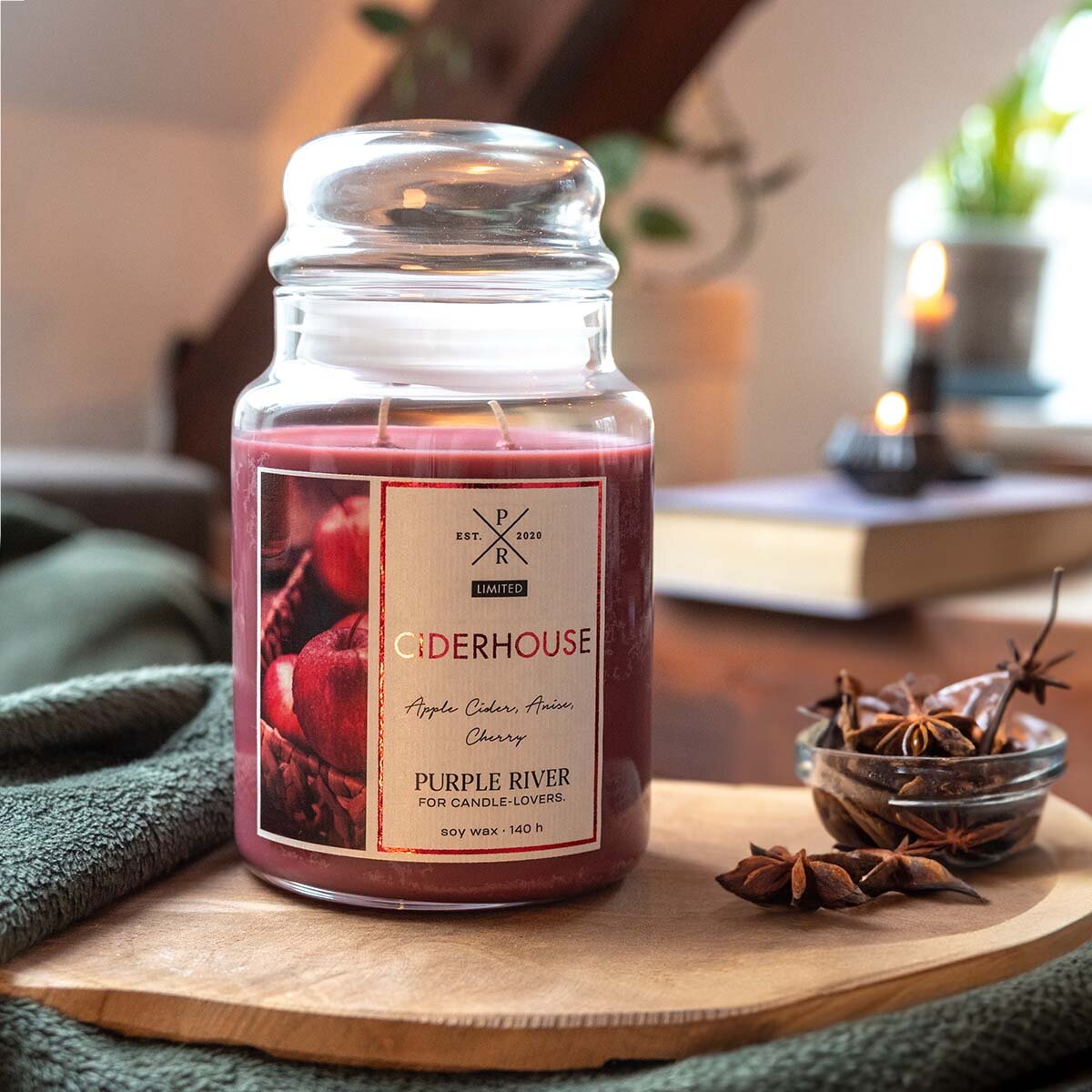Nachhaltige Premium-Duftkerze von Purple River mit extralanger 140h-Brenndauer. Hochwertige Deko im Glas. Made in Germany - inszeniert auf Holzbrett neben Nelken & Buch im Wohnzimmer.