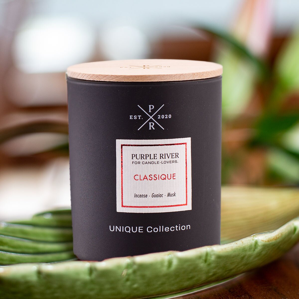 Nachhaltige Premium-Duftkerze von Purple River in schwarz mit extralanger 140h-Brenndauer. Hochwertige Wellness-Deko im Glas und mit Holzdeckel. Made in Germany - inszeniert auf grüner Keramikschale  im Wellnessbereich.