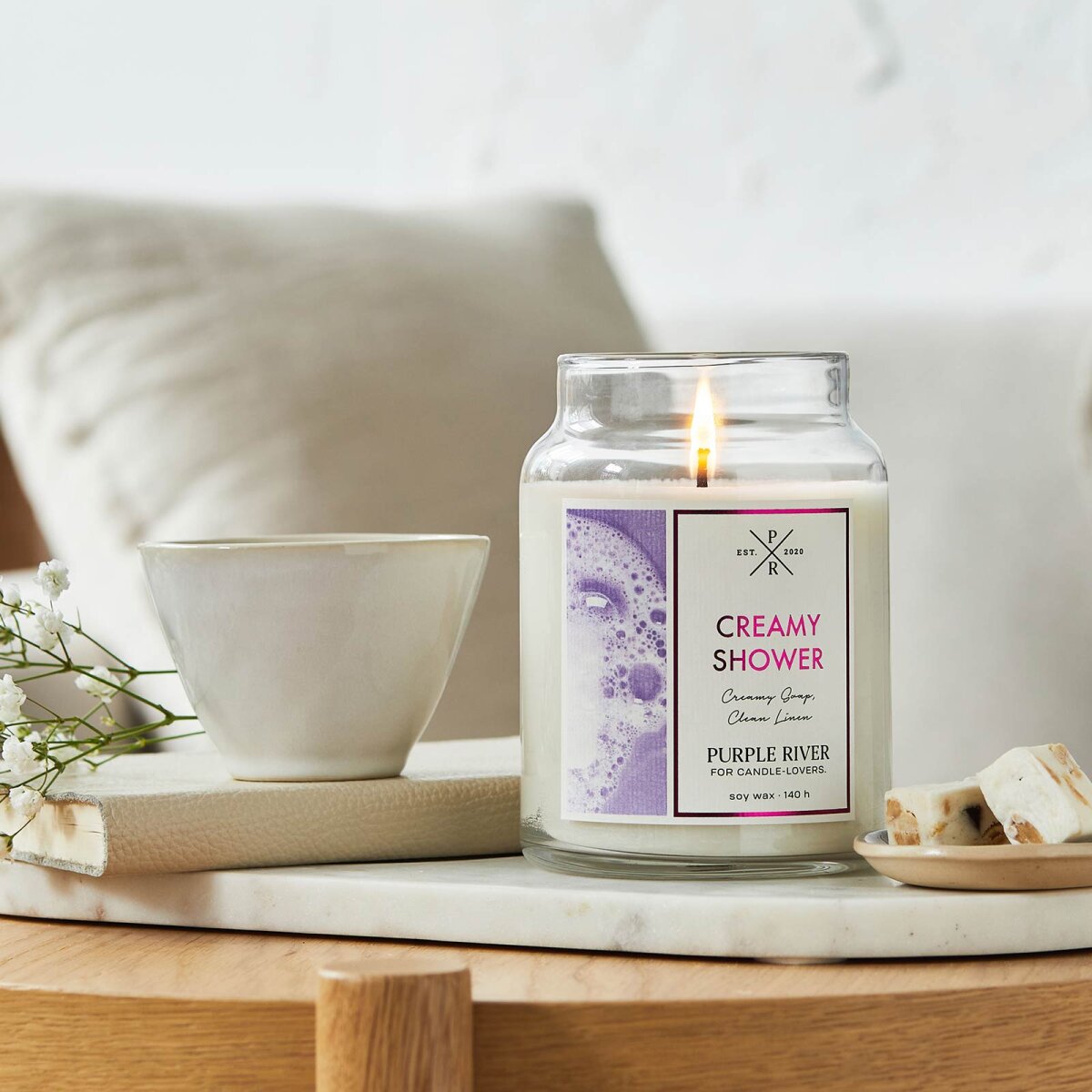 Nachhaltige Premium-Duftkerze von Purple River mit extralanger 140h-Brenndauer. Hochwertige Wellness-Deko im Glas. Made in Germany - inszeniert auf Steinplatte neben Pralinen & Buch im Wohnzimmer.