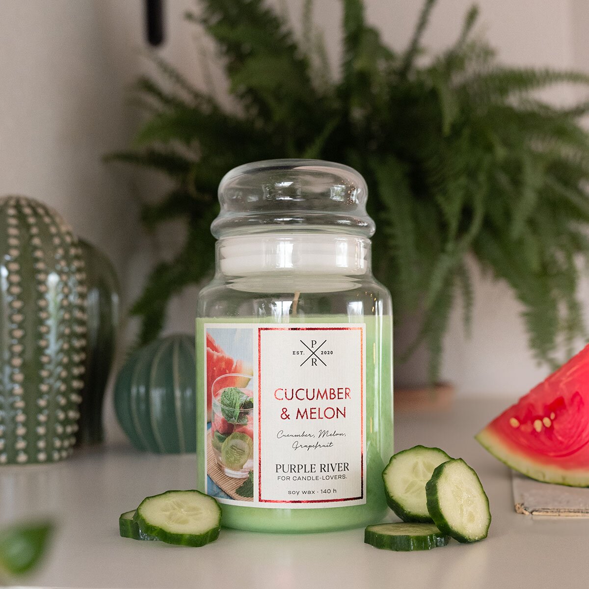Nachhaltige Premium-Duftkerze von Purple River mit extralanger 140h-Brenndauer. Hochwertige Wellness-Deko im Glas. Made in Germany - inszeniert auf Tisch neben Gurke & Melone im Wellnessbereich.