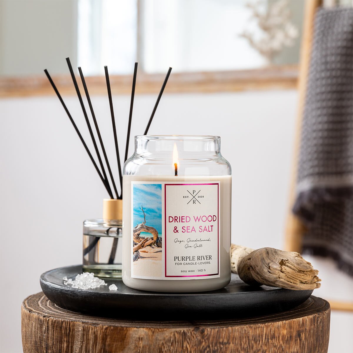 Nachhaltige Premium-Duftkerze von Purple River mit extralanger 140h-Brenndauer. Hochwertige Wellness-Deko im Glas. Made in Germany - inszeniert auf Keramikteller neben Salz, Todholz & Diffuser im Badezimmer.