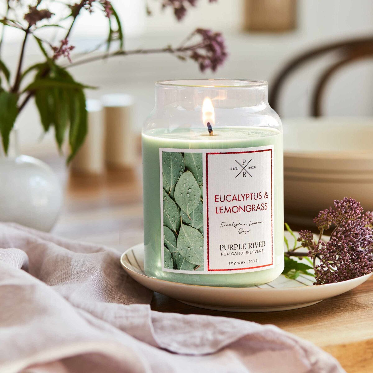 Nachhaltige Premium-Duftkerze von Purple River mit extralanger 140h-Brenndauer. Hochwertige Wellness-Deko im Glas. Made in Germany - inszeniert auf Holztisch neben Pflanzen & Küchentuch im Wohnzimmer.