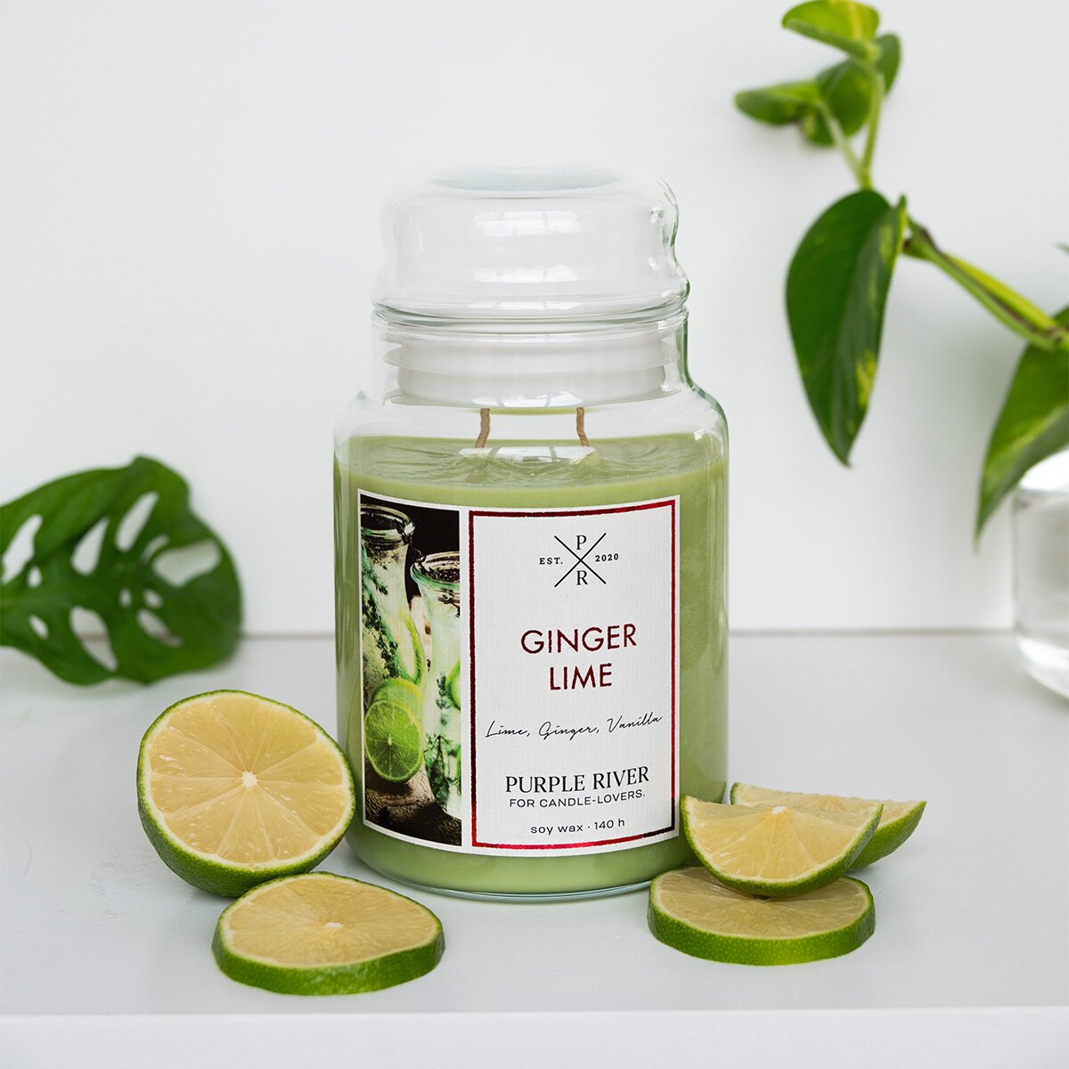 Nachhaltige Premium-Duftkerze von Purple River mit extralanger 140h-Brenndauer. Hochwertige Wellness-Deko im Glas. Made in Germany - inszeniert auf Tisch neben Limetten & Grünpflanzen im Wellnessbereich.