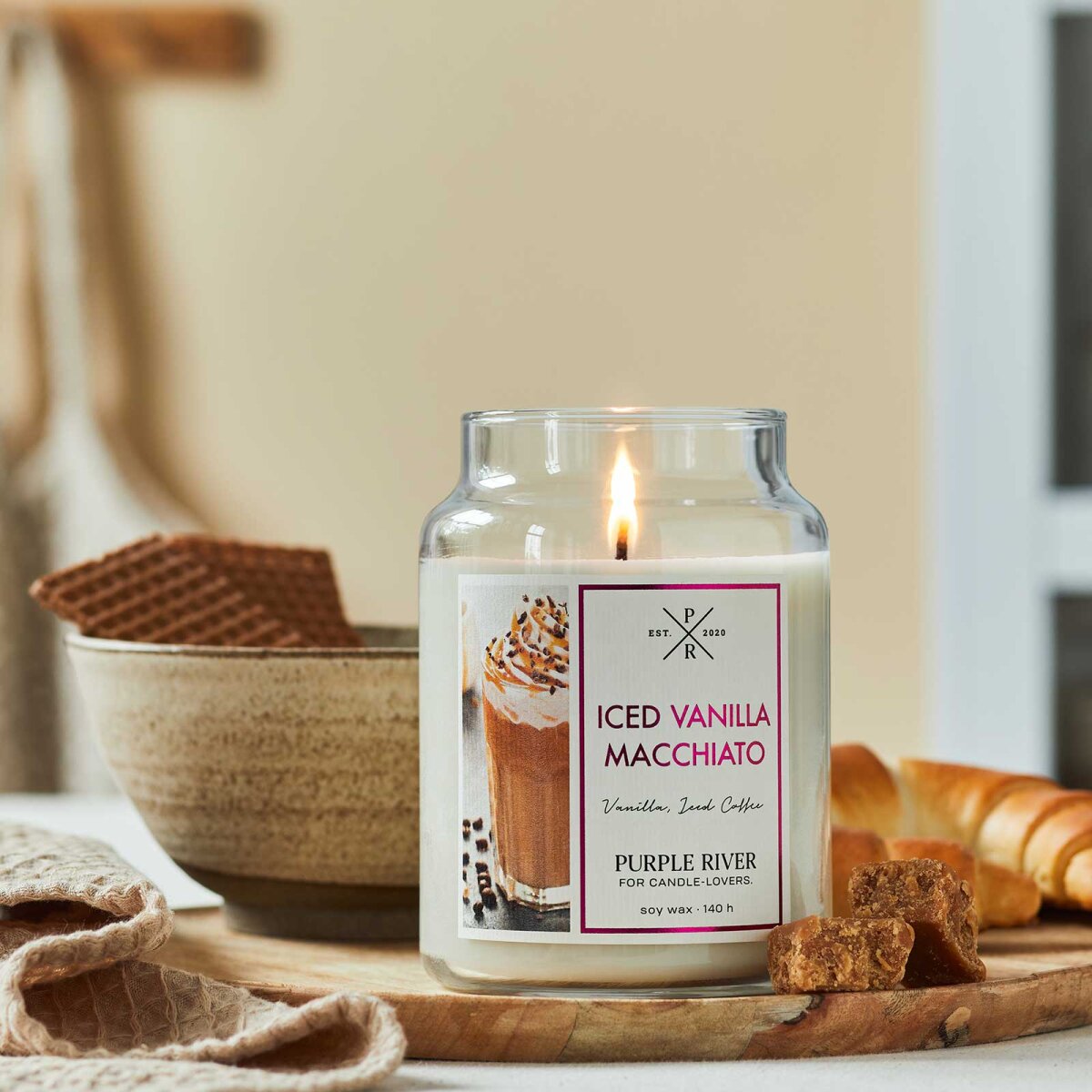 Nachhaltige Premium-Duftkerze von Purple River mit extralanger 140h-Brenndauer. Hochwertige Wellness-Deko im Glas. Made in Germany - inszeniert auf Holzbrett neben süßem Gebäck & in Küche.