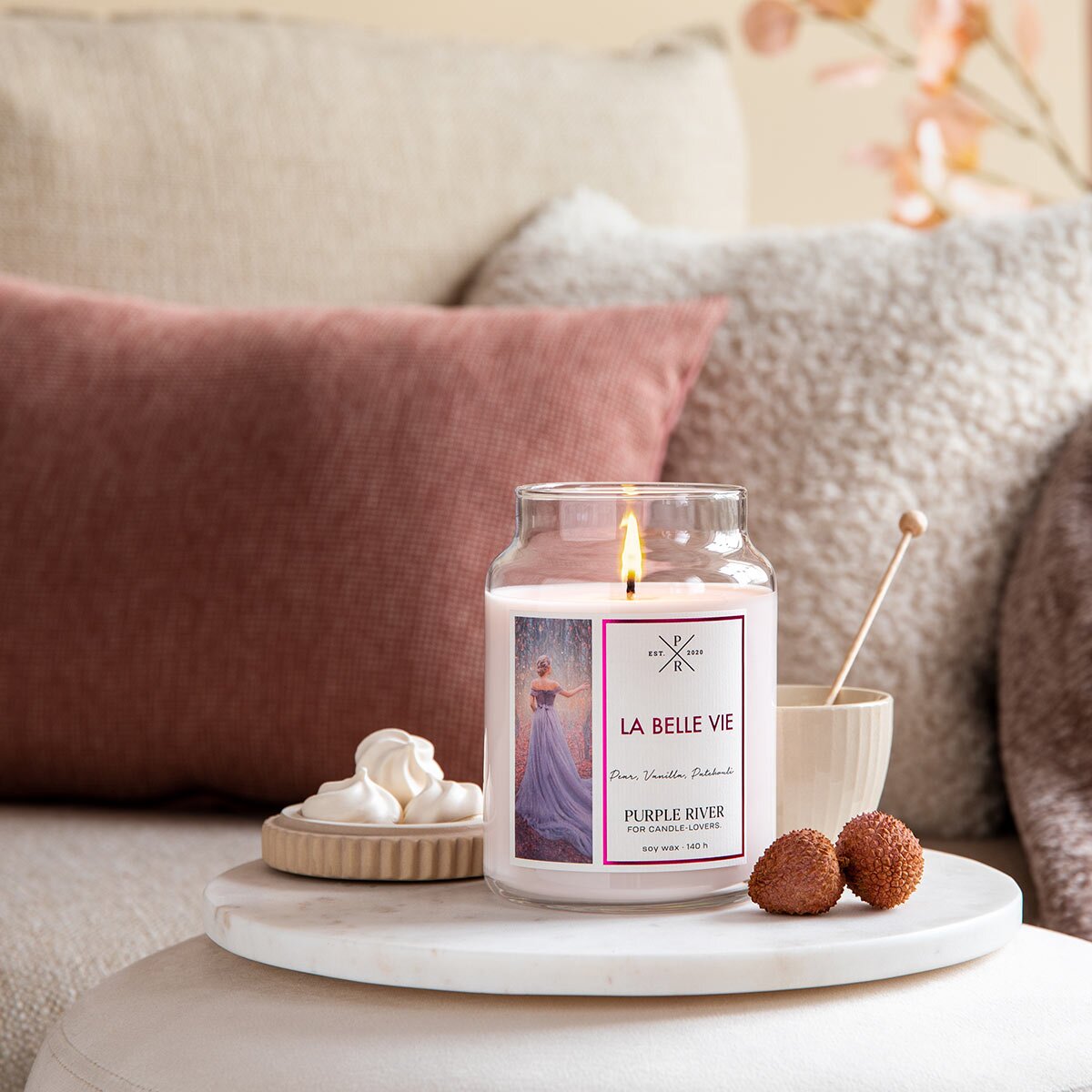 Nachhaltige Premium-Duftkerze von Purple River mit extralanger 140h-Brenndauer. Hochwertige Wellness-Deko im Glas. Made in Germany - inszeniert auf Steinplatte neben Tee & Süßigkeiten im Wohnzimmer.