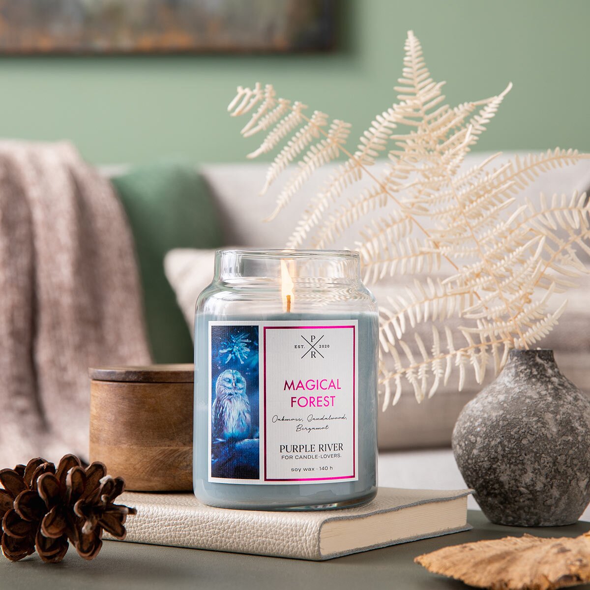 Nachhaltige Premium-Duftkerze von Purple River mit extralanger 140h-Brenndauer. Hochwertige Wellness-Deko im Glas. Made in Germany - inszeniert auf Buch neben Pflanzendeko & Tannenzapfen im Wohnzimmer.