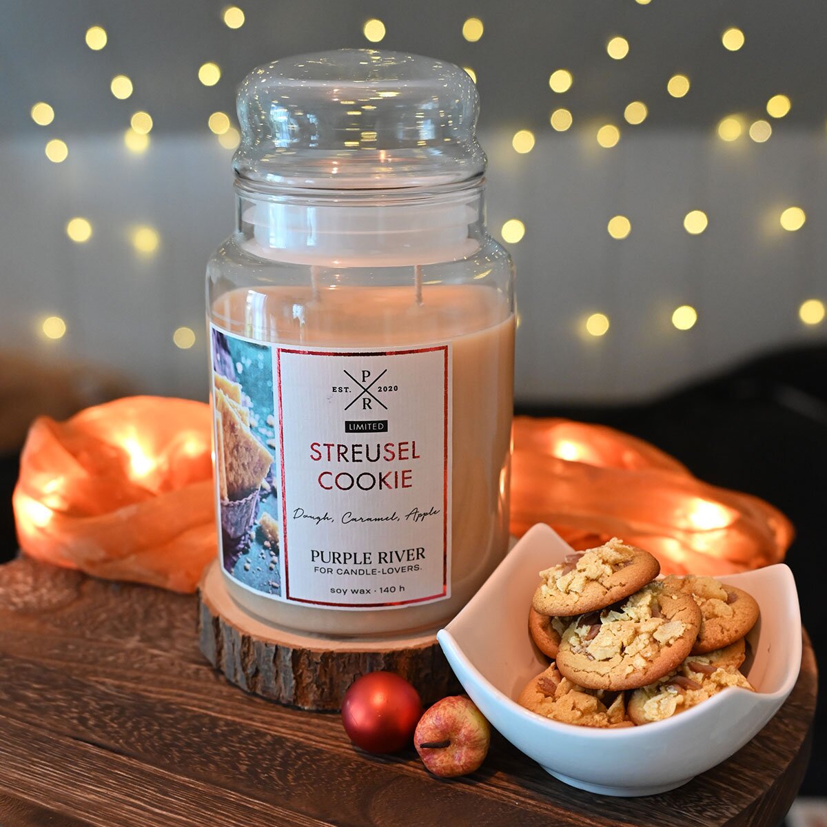 Nachhaltige Premium-Duftkerze von Purple River mit extralanger 140h-Brenndauer. Hochwertiges Geschenk im Glas. Made in Germany - inszeniert auf Holztisch neben Plätzchen & Lichterkette auf Geschenketisch.