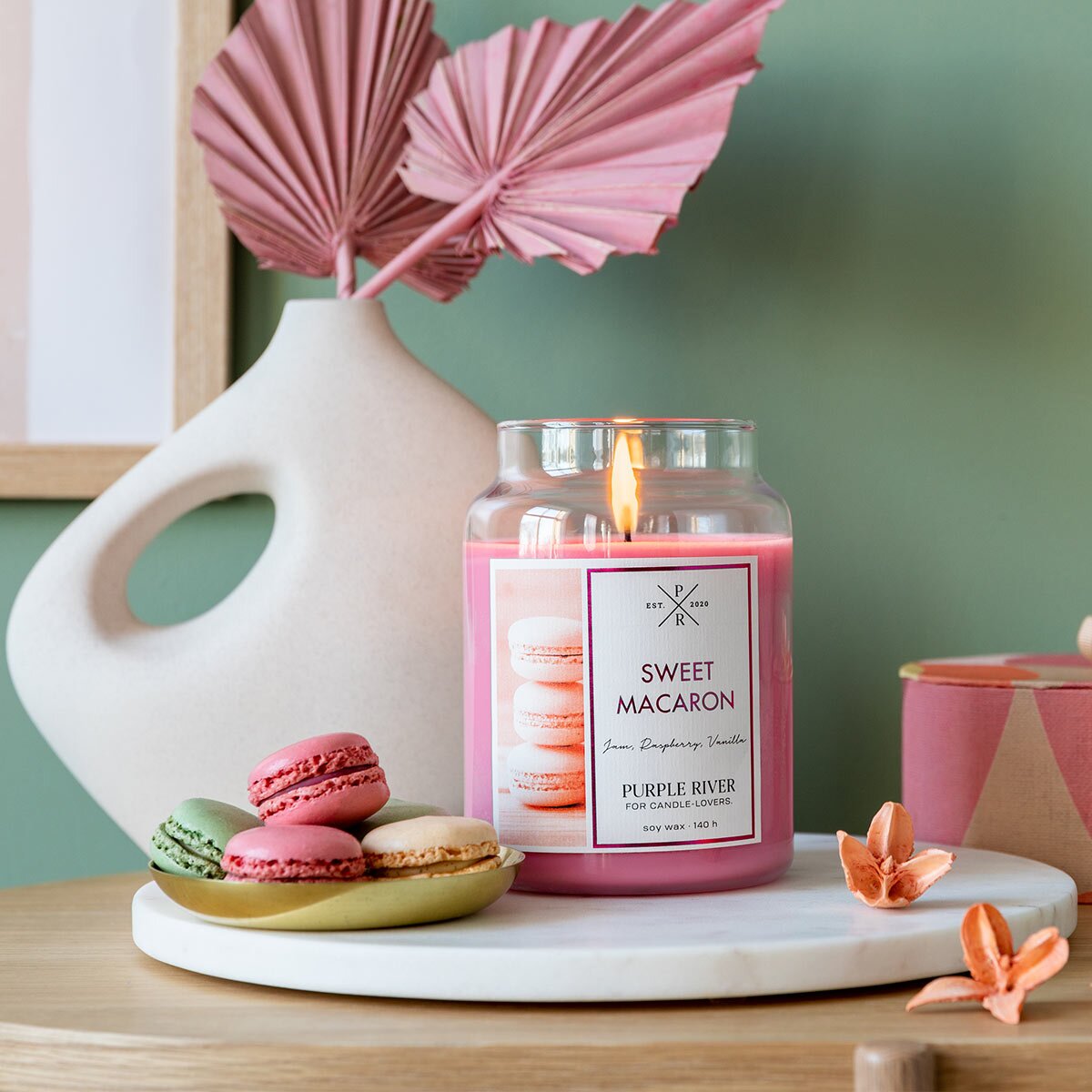 Nachhaltige Premium-Duftkerze von Purple River mit extralanger 140h-Brenndauer. Hochwertige Geschenkidee. Made in Germany - inszeniert auf Holztisch neben Macarons & Bucheckern im Wohnzimmer.
