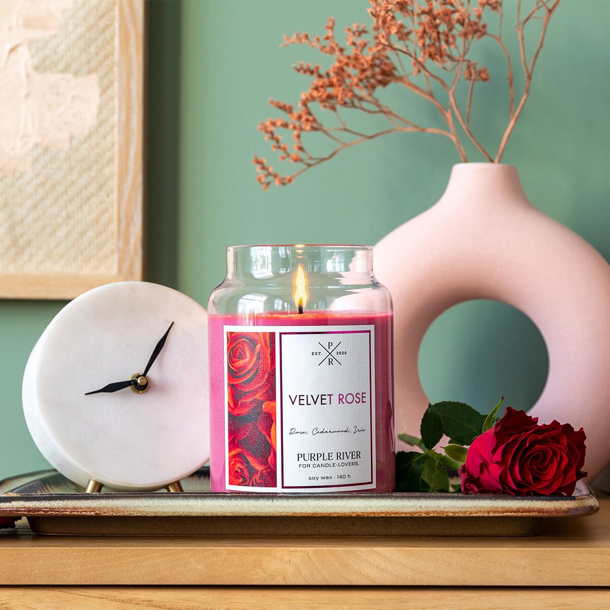 Nachhaltige Premium-Duftkerze von Purple River mit extralanger 140h-Brenndauer. Hochwertige Wellness-Deko im Glas. Made in Germany - inszeniert auf Holzbrett neben Rose & Uhr im Flur.
